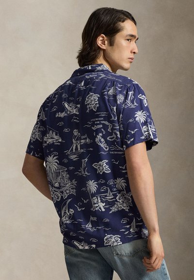 Młody mężczyzna stojący bokiem, ubrany w granatową koszulę z krótkim rękawem z białymi tropikalnymi i marynistycznymi wzorami oraz jasnoniebieskie dżinsy.