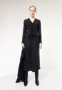 Top nero a maniche lunghe con arricciature sul davanti, abbinato a una gonna nera coordinata. La modella indossa stivali neri lucidi fino al ginocchio e tiene in mano un blazer.