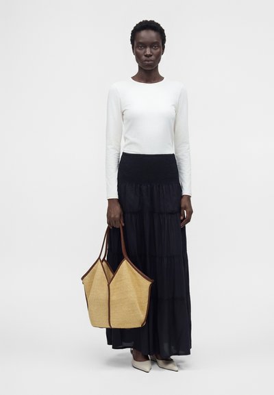 Femme portant un haut blanc à manches longues et une jupe maxi noire à volants, tenant un grand sac fourre-tout beige avec des poignées marron, debout sur un fond uni.