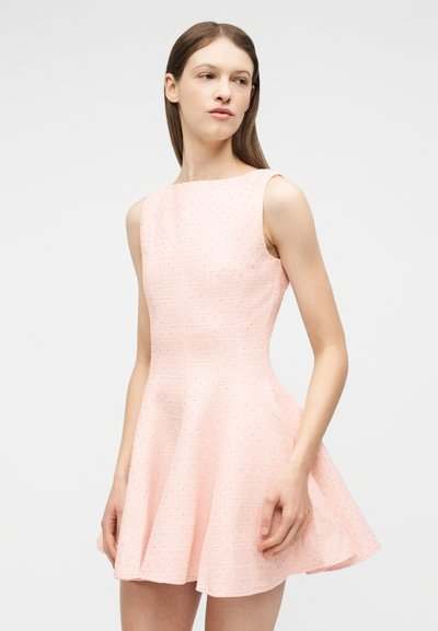 Νεαρή γυναίκα που φοράει αμάνικο, ανοιχτό ροζ, υφασμάτινο φόρεμα με καμπάνα, στέκεται με το κεφάλι ελαφρώς γυρισμένο στο πλάι μπροστά από απλό φόντο.