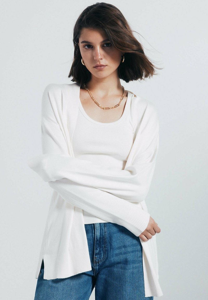 Bílý cardigan s ribbovanými manžetami a lemem, vrstvený přes bílý tílko. Nosený s modrými džíny, které mají hladkou a jemnou texturu.