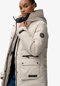 Beżowa ocieplana parka z wysokim kołnierzem, zapięciem na zamek błyskawiczny, czarnymi akcentami i wieloma kieszeniami. Posiada naszywkę z logo na rękawie.