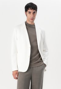Blazer bianco su misura in tessuto morbido, chiusura con un bottone e rever a punta, abbinato a un top marrone fitted e pantaloni grigi.