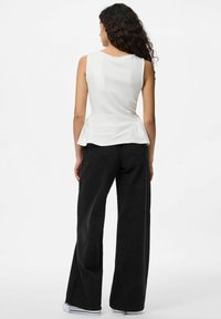Una persona con lunghi capelli ricci indossa un top peplum bianco e pantaloni neri a gamba larga, vista di spalle su uno sfondo chiaro.