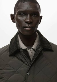 Giacca trapuntata verde scuro con colletto in velluto a coste, con chiusura frontale a zip e una camicia grigia testurizzata sotto.