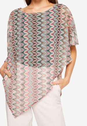 Kobieta ubrana w luźny, asymetryczny top z krótkim rękawem w wielokolorowy wzór w zygzag, białe spodnie, ręce w kieszeniach.