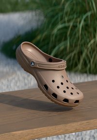 Sabots marron clair avec des trous de ventilation, semelle épaisse et sangle texturée, fabriqués en matériau plastique lisse. Présente un emblème de la marque sur le côté.