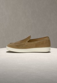 Chaussure en daim marron clair sans lacets avec semelle en caoutchouc blanche, présentée sur une surface gris clair sur fond beige.