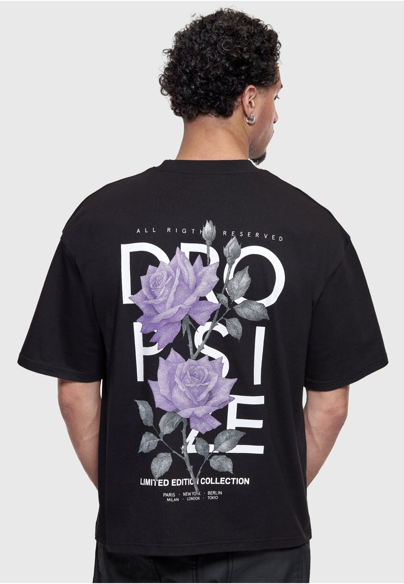 Homme portant un T-shirt noir oversize avec de grandes roses violettes et un texte blanc indiquant « DROP SIDE » et « LIMITED EDITION COLLECTION » dans le dos.