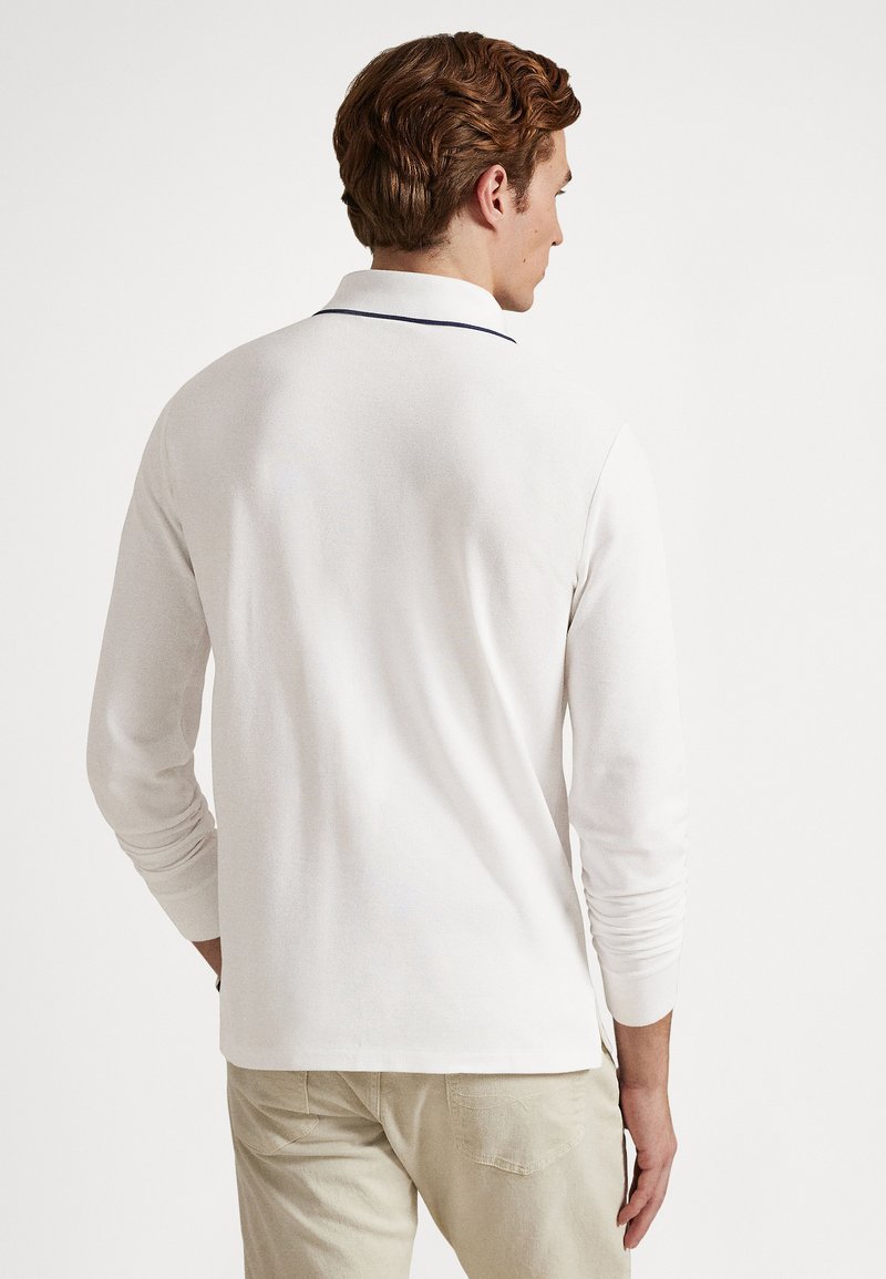 Homme de dos portant un polo à manches longues blanc avec un bord sombre au col et un pantalon beige clair.