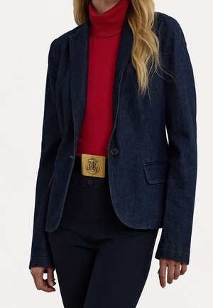 Femme portant un blazer en denim foncé, un col roulé rouge, un pantalon noir et une boucle de ceinture dorée avec un logo ancre et initiales.