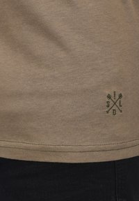 Kaki kortærmet t-shirt lavet af bomuld med en subtil tekstur. Har et grønt broderet logo med krydsspile ved sømmen.