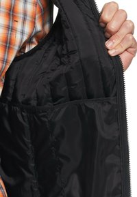 Schwarze isolierte Jacke mit gesteppter Innenseite, ausgestattet mit einem Reißverschluss und einer Tasche. Im Hintergrund ist ein kariertes Hemd zu sehen.