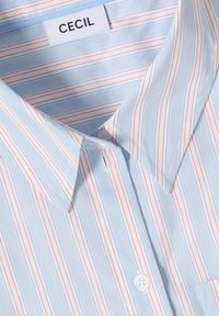 Chemise bleu clair boutonnée avec des rayures verticales bleues et rouges. Elle comporte un col pointu et une étiquette marquée "CECIL" à l'intérieur du col.