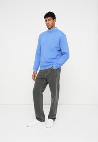 Svetlomodrá mikina s rebrovanými manžetami, preložená cez kockovanú košeľu s golierom, v kombinácii s tmavosivými džínsami a bielymi teniskami.