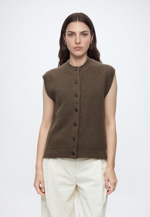 Femme aux longs cheveux bruns portant un gilet en tricot marron sans manches boutonné et un pantalon couleur crème, debout devant un fond blanc uni.