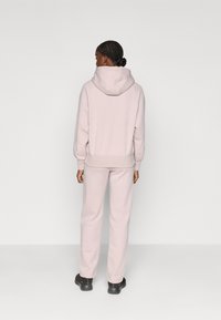 Haut à capuche rose pâle avec une large capuche et des poignets côtelés, associé à un pantalon de jogging assorti. Texture douce et coupe décontractée. Chaussures noires visibles.