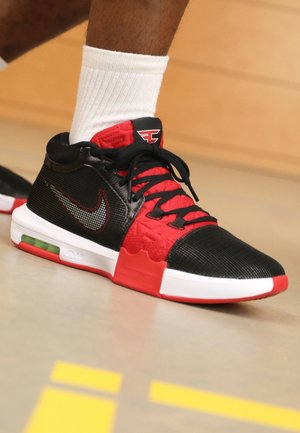 Basket Nike rouge et noire avec semelle à air visible et lacets noirs, portée avec une chaussette blanche haute sur un sol intérieur avec des marquages jaunes.