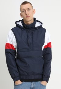 Marineblå pullover-jakke med hvide og røde accenter, mellem høj krave, frontlomme, snoretræk i hætten og elastiske manchetter og kant.