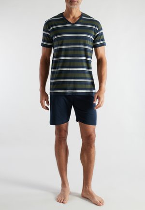 Man wearing navy blue shorts and a green, navy, and white striped short-sleeve V-neck shirt, standing barefoot against a plain background.