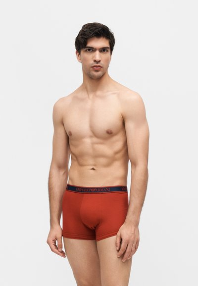 Άνδρας χωρίς πουκάμισο που φοράει σορτς Emporio Armani σε σκουριασμένο χρώμα, στέκεται μπροστά σε ένα απλό ανοιχτό γκρι φόντο.