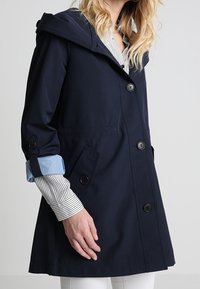 Chaqueta con capucha azul marino de corte entallado, con cierre de botones, dos bolsillos laterales y detalles en azul claro en los puños.