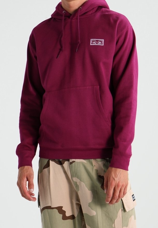 Homme portant un sweat à capuche bordeaux avec un petit logo sur la poitrine et un pantalon cargo camouflage beige et vert.