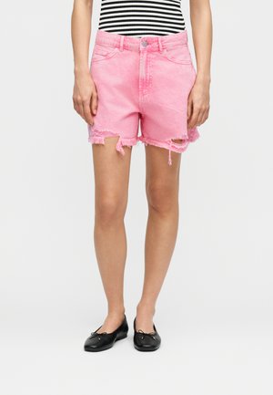 Γυναίκα που φοράει ροζ σκισμένο τζιν σορτς, μαύρα μπαλαρίνια και ριγέ μαύρο-λευκό τοπ, στέκεται μπροστά σε απλό ανοιχτό φόντο.