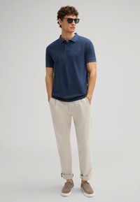 Granatowa koszulka polo z krótkim rękawem, zestawiona z jasnobeżowymi spodniami. Model nosi ciemne okulary przeciwsłoneczne i szare buty typu slip-on w wzory.