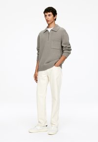 Grau gestrickter Pullover mit Reißverschlusskragen und Fronttasche, kombiniert mit cremefarbener Hose und weißen Sneakers. Schlichtes und modernes Design.