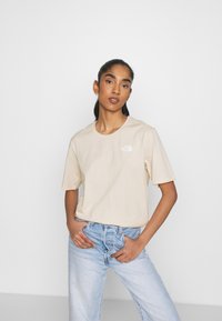 T-shirt beige a maniche corte in cotone, con un logo bianco sul lato sinistro del petto. Abbinato a jeans a gamba larga di un blu chiaro.