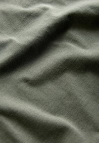 Olīvu zaļš audums ar mīkstu, tekstūru virsmu, kas parāda vieglas krokas un gludu, vienmērīgu audumu visā materiālā.