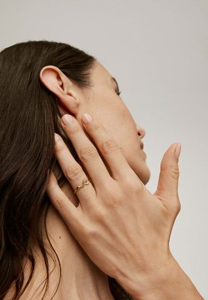 Mujer con cabello largo y oscuro que toca su cuello, llevando un delicado anillo de oro con un diseño retorcido en su dedo anular.