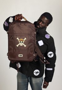 Sac à dos en nylon marron avec un design de tête de mort et de os croisés. Il présente une forme rectangulaire, des bretelles ajustables et une poignée sur le dessus.