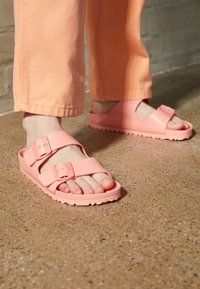 Chanclas de goma rosas con doble tira ajustable y una suela texturizada, llevadas con pantalones naranja claro sobre una superficie lisa.