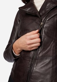 Bruine leren jas met een hoge kraag, ritssluiting en een zachte textuur. Close-up van een hand die de rits vasthoudt.