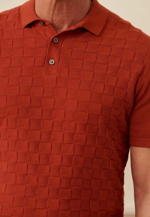 Polo de manga corta en rojo óxido, con un patrón texturizado de cuadros, tres botones marrones en el cuello y un material de punto suave.