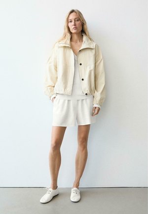 Kobieta o długich blond włosach stoi na tle białej ściany, ubrana w jasnobeżową kurtkę, sweter, szorty oraz białe buty na sznurowadła.