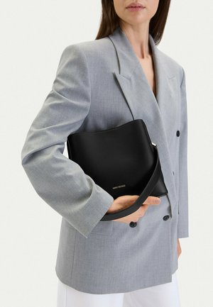 Kobieta ubrana w jasnoszary blezer, trzymająca czarną torebkę Gino Rossi na jednolitym białym tle.