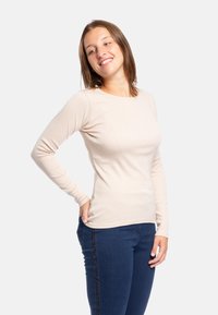 Maglia beige a maniche lunghe in tessuto morbido, con scollatura tonda e silhouette aderente, abbinata a jeans blu scuro con decorazioni.