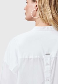 Weißes Baumwollhemd mit Stehkragen, Knopfverschluss am Rücken und kleinem Markenetikett am Hals. Glatte Textur, oversized Schnitt.