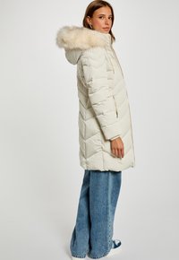 Pufferjakke i creme med en pelsforet hætte, quiltet tekstur og lynlås lukning. Parret med brede blå jeans og sneakers.