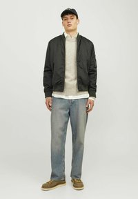 Sort bomberjakke over en beige striktrøje, hvid skjorte og blå jeans. Brune sko med gule snørebånd og en sort kasket.