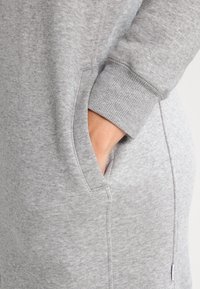 Grauer Sweatshirt mit einer weichen Textur, ausgestattet mit einer geräumigen Seitentasche und gerippten Bündchen. Der Stoff wirkt leicht strukturiert und langlebig.