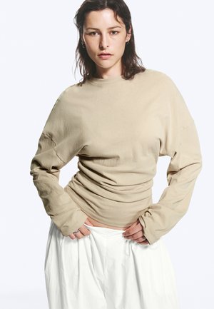 Femme aux cheveux foncés portant un haut beige à manches longues et un pantalon blanc, regardant la caméra, les mains près de la taille, sur un fond uni.