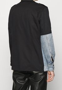 Černá a džínová bunda se strukturovaným siluetem. Černá bavlněná část, džínové rukávy a postranní rozparky s akcenty knoflíků.