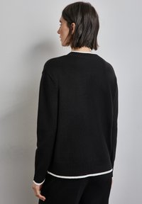 Svart tröja med avslappnad passform, ribbad krage och fåll, med en vit randig detalj vid ärmsluten och ett lagerutseende i nederkant.