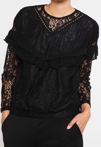 Top en dentelle noire avec des manches longues, des accents de volants superposés, un décolleté en V et des motifs floraux intricats. Tissu texturé avec une coupe décontractée.