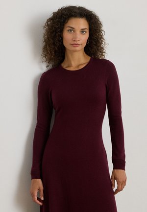 Frau mit lockigem Haar, die ein langärmliges, tailliertes, dunkelburgunderfarbenes Kleid trägt und vor einer hellgrauen Wand steht.