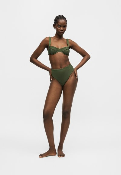 Femme debout pieds nus portant un bikini vert olive avec un bas taille haute et un haut structuré, posant avec les mains sur les hanches devant un fond blanc.